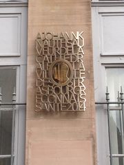 un profil en bronze doré entouré des lettres du texte réfléchit la cathédrale. Se trouve à gauche de l' entrée de la poste.